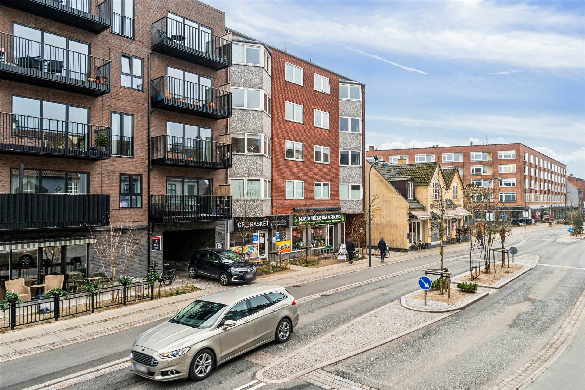 To biler kører langs en vej med søjler og gavlhuse, træer, grønne buske og terrasser