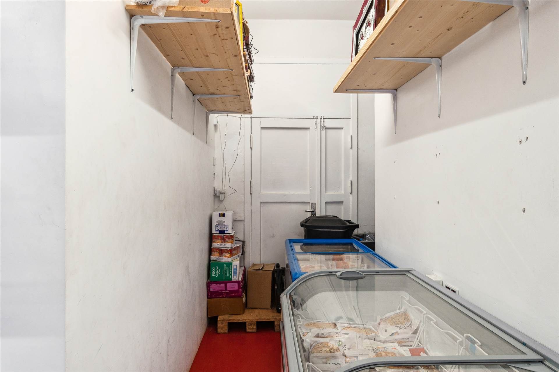 To små frysere med flere krydsfinerkasser og bokse og en sort skraldespand i et køkken med hvide vægge og rødt gulv