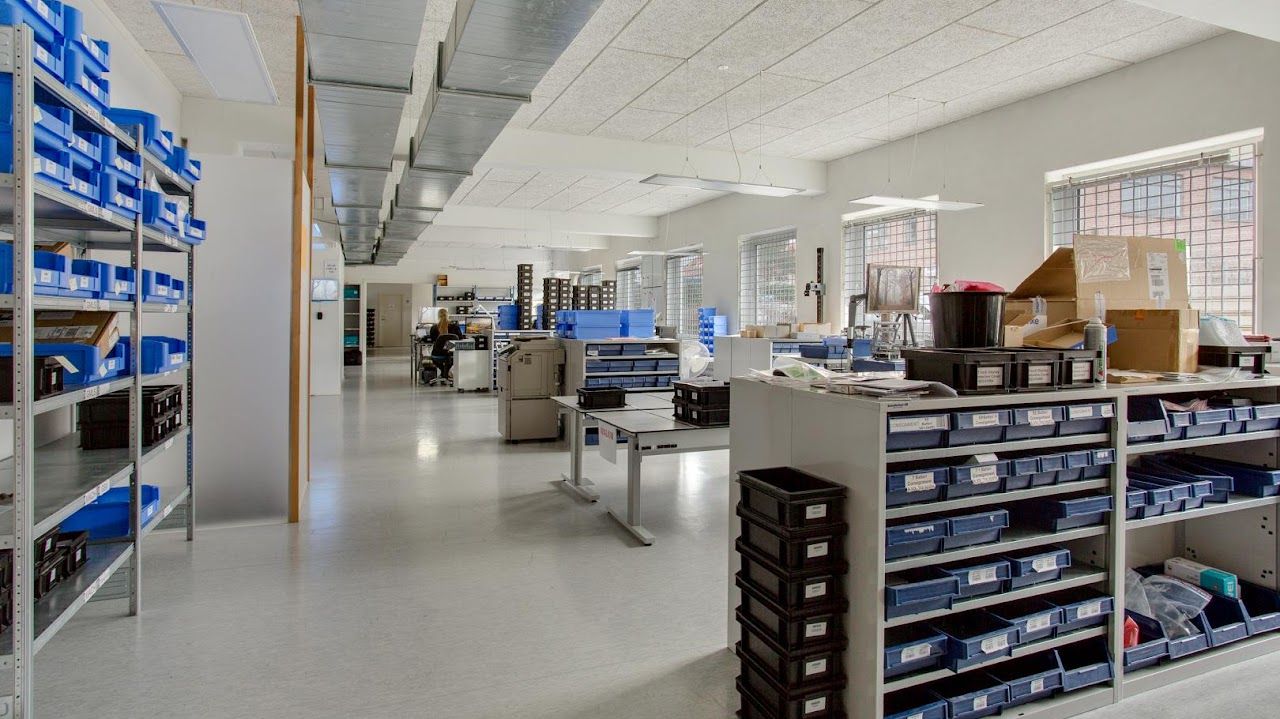 Lager med høje hylder og bokse. Sorte og blå bokse står på hylderne. Vinduer med stålrammer og ruller. Hvide lofter med