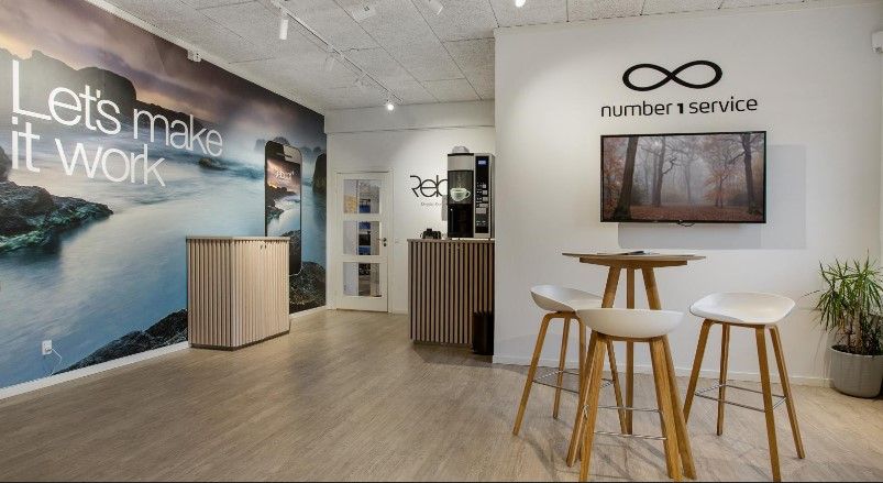 Kontor med borde og stole samt en TV-skærm og en kaffemaskine på væggen. Der er en stor billedtapet med teksten "Let's make