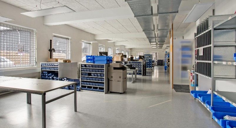 Et rum med vægge, hvor der er flere hylder med blå kasser og en printer i midten, samt et bord med metalben og et gulv med