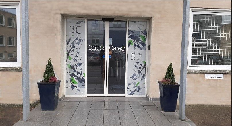 Et moderne kontorbyggeri med store glasdøre med mærket Careo Careo, to plantepotter foran og en person i spejlet