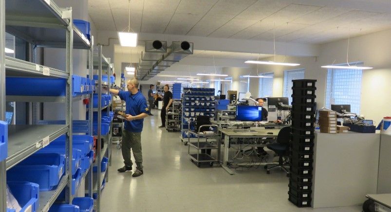 Man i blåt tøj står ved et stort hyldesystem og vender sig mod kameraet i et stort lokale med flere medarbejdere og hylder