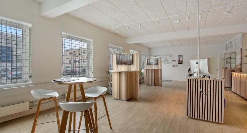 Et moderne kontor med højt loft, store vinduer, og en minimalistisk indretning med lyse trægulve og hvide vægge. Der er