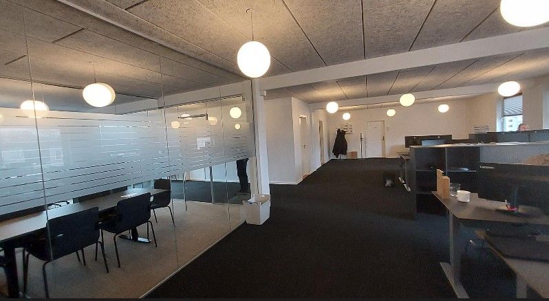 Moderne kontor med glasvægge og lyse loftslampe. Bord og stole samt en person i en jakke står i rummet.