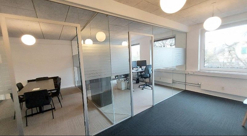 Lyst og moderne kontor med glasvægge og stolpefordelt belysning i hvid og grå. Komplet møbleret med bord, stol og computer.