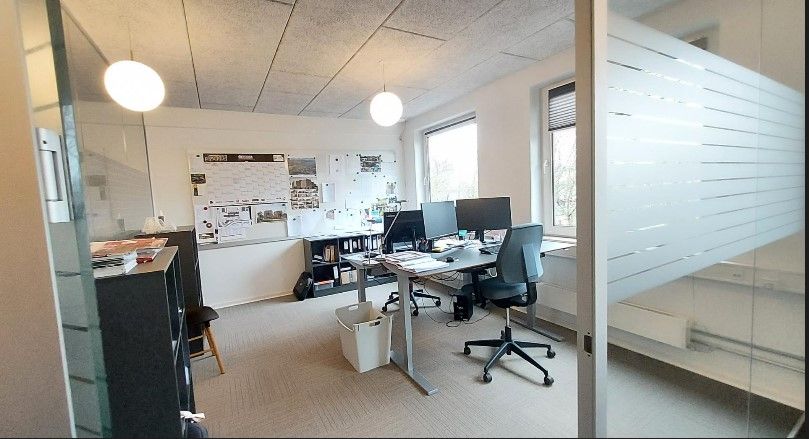 Kontor med to borde, hver med to skærme og en skrivemaskine, samt to stolpe, en skraldespand og en lysekroner