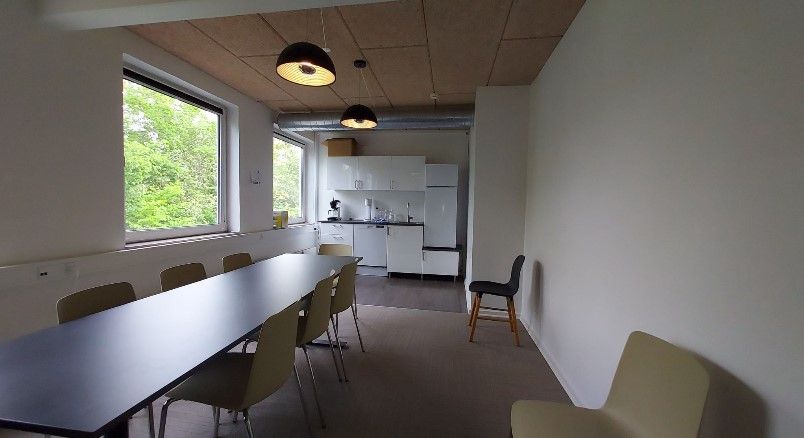 Lyst, nyistandsat kontor med lyse loftslamper, langt borde, stole, køkken, hvidt køleskab og vindue til udendørsarealet.