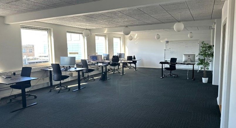 Kontorrum med skriveborde, computere og lysekroner. I baggrunden ser man vinduer med byudsigt. Hjørne med plante og