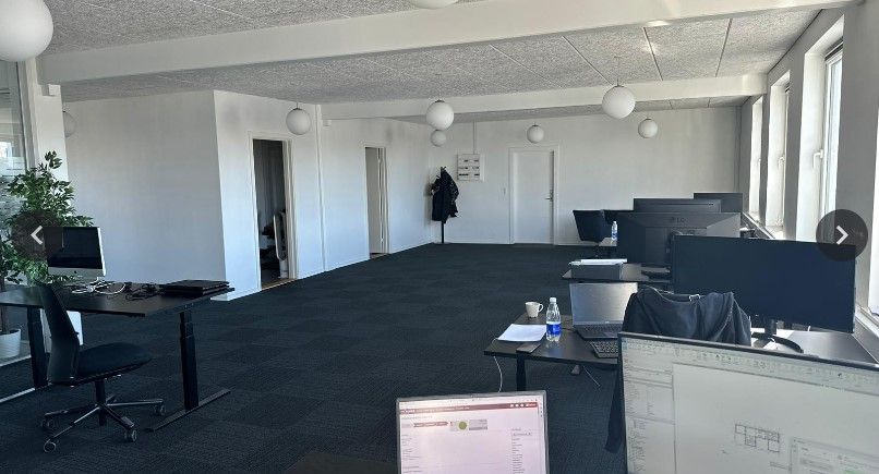 Lyst og moderne kontor med flere computere, skriveborde, stole og lyse gulve samt store vinduer.