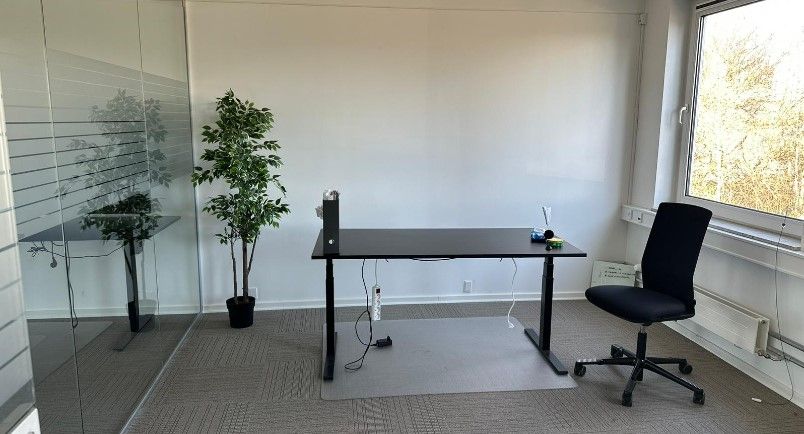 Et tomt kontor med glasvægge og gulvtæppe. Der er en sort skrivebord og en sort stol. På skrivebordet er der en computer og