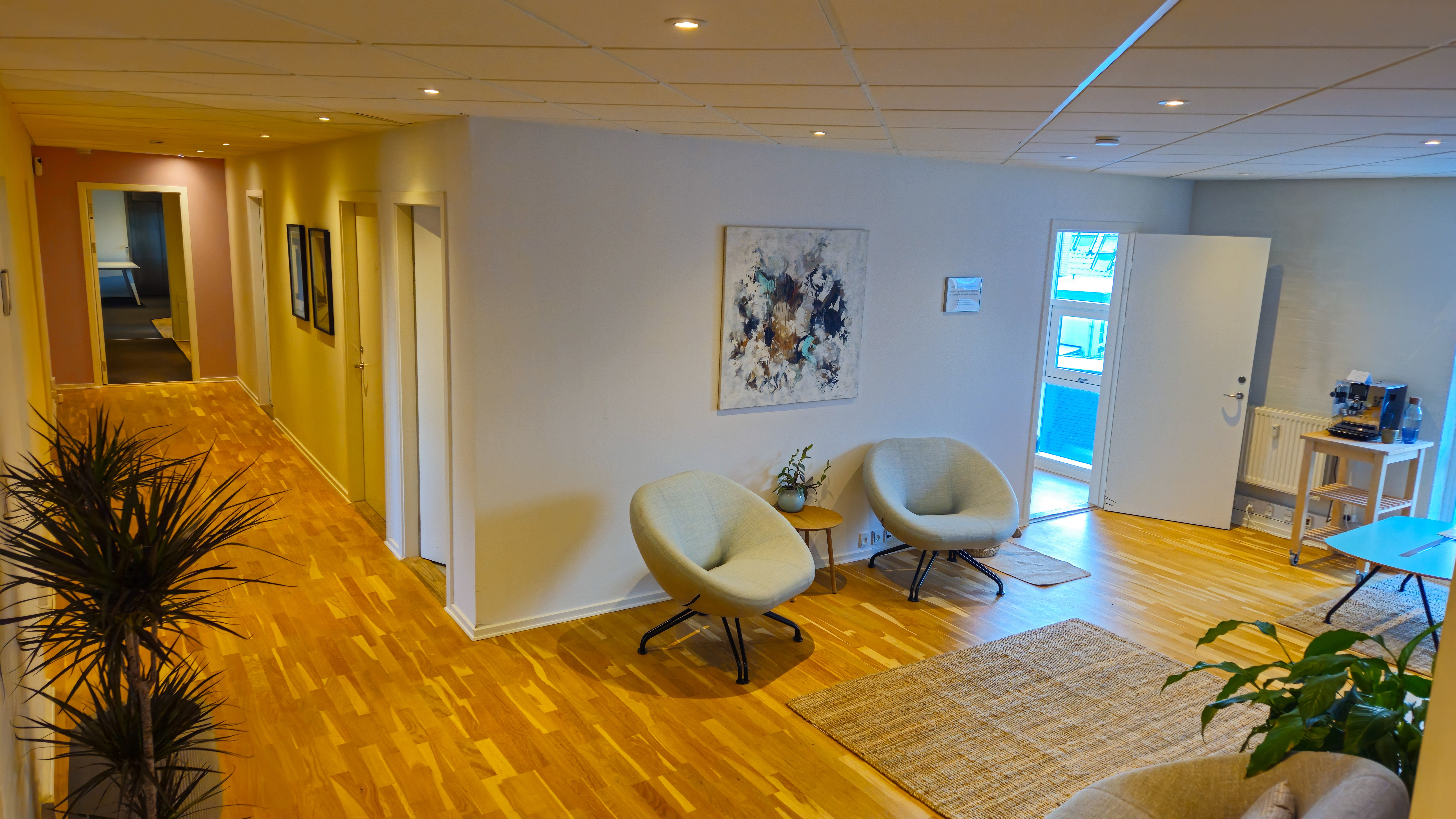 Moderne indretning med to lyse sædestole og en plante ved en hjørnevæg med maleri og sammenhængende døre