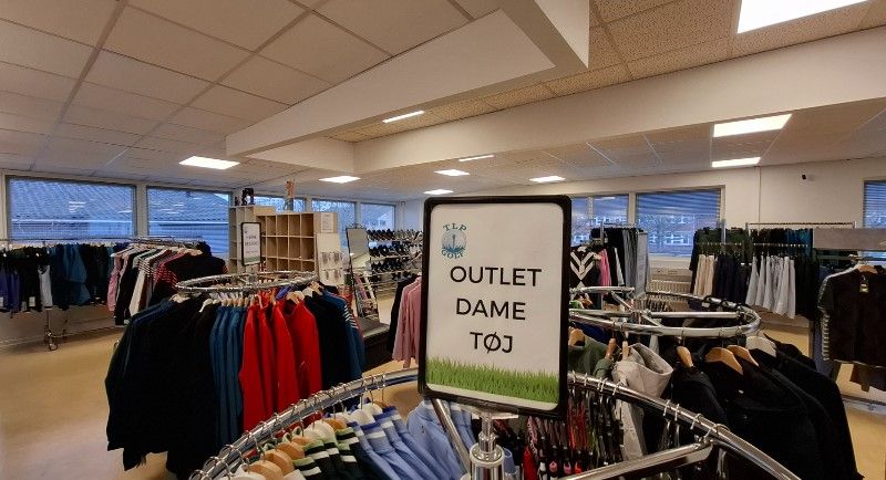 En klædeskabende med diverse tøj i sort, rødt, gult og blåt. Der er skilte med teksten Outlet Dame Tøj. Der er vinduer i