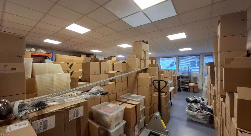 Lager med store mængder papkasser, plastmaterialer, og emballage. Langt væk er der en læsserampe og vinduer med udsigt til