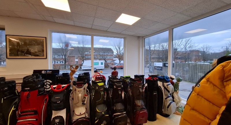 Golfbutik med udstille klubsæt og golftasker, udsigt ud i en bygade med biler og skov, og en mand i golftøj og hætte står på