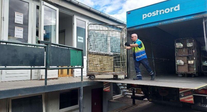En mand i sikkerhedsvest og handsker ligger og håndterer en kæde, der er lastet på en lastbil med Postnord-mærkning. Kæden