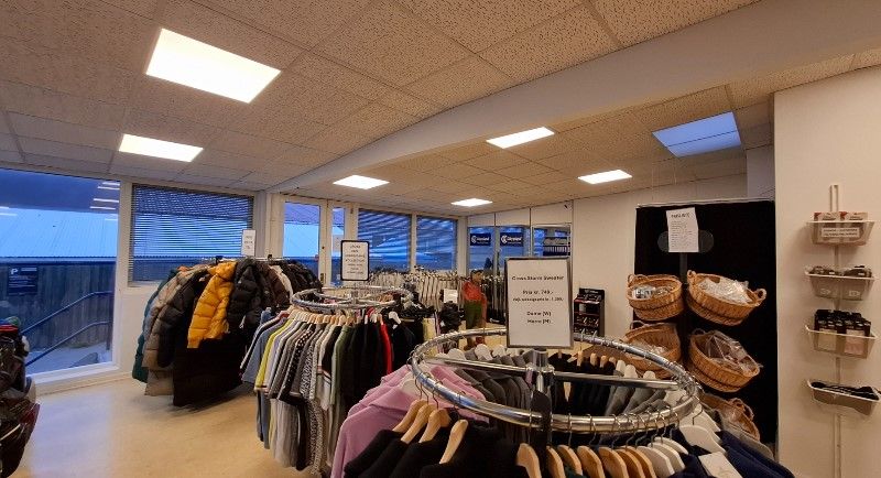 Butik med klæder og jakker på stativer og vægge, sammen med kurve og hylder med tøj og andre produkter, vinduer med udsigt