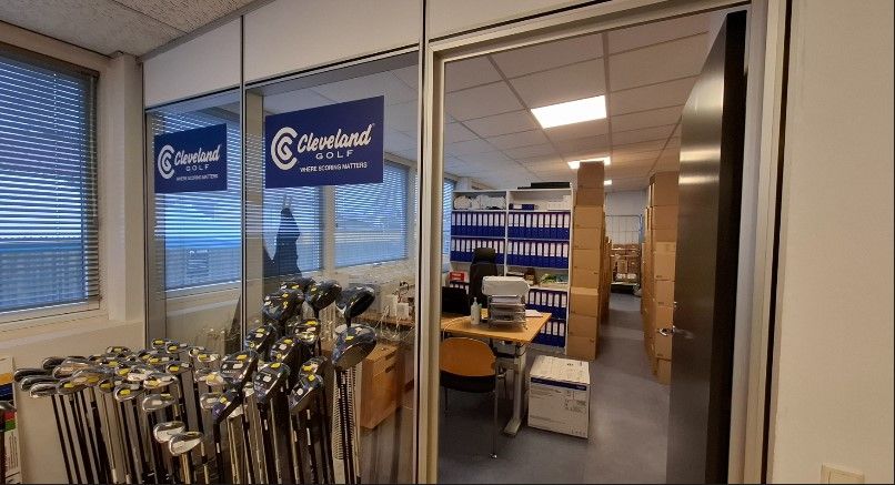 Golfklubber og udstyr står opstillet i en stor butik. Der er et skriftligt skilt på væggen med Cleveland Golf. Der er flere