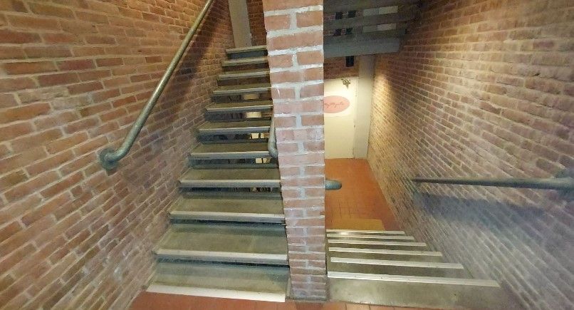 En stejl trappe i røde mursten fører ned i et underliggende rum. Der er to rækker trappetrin med håndtag og et lys.