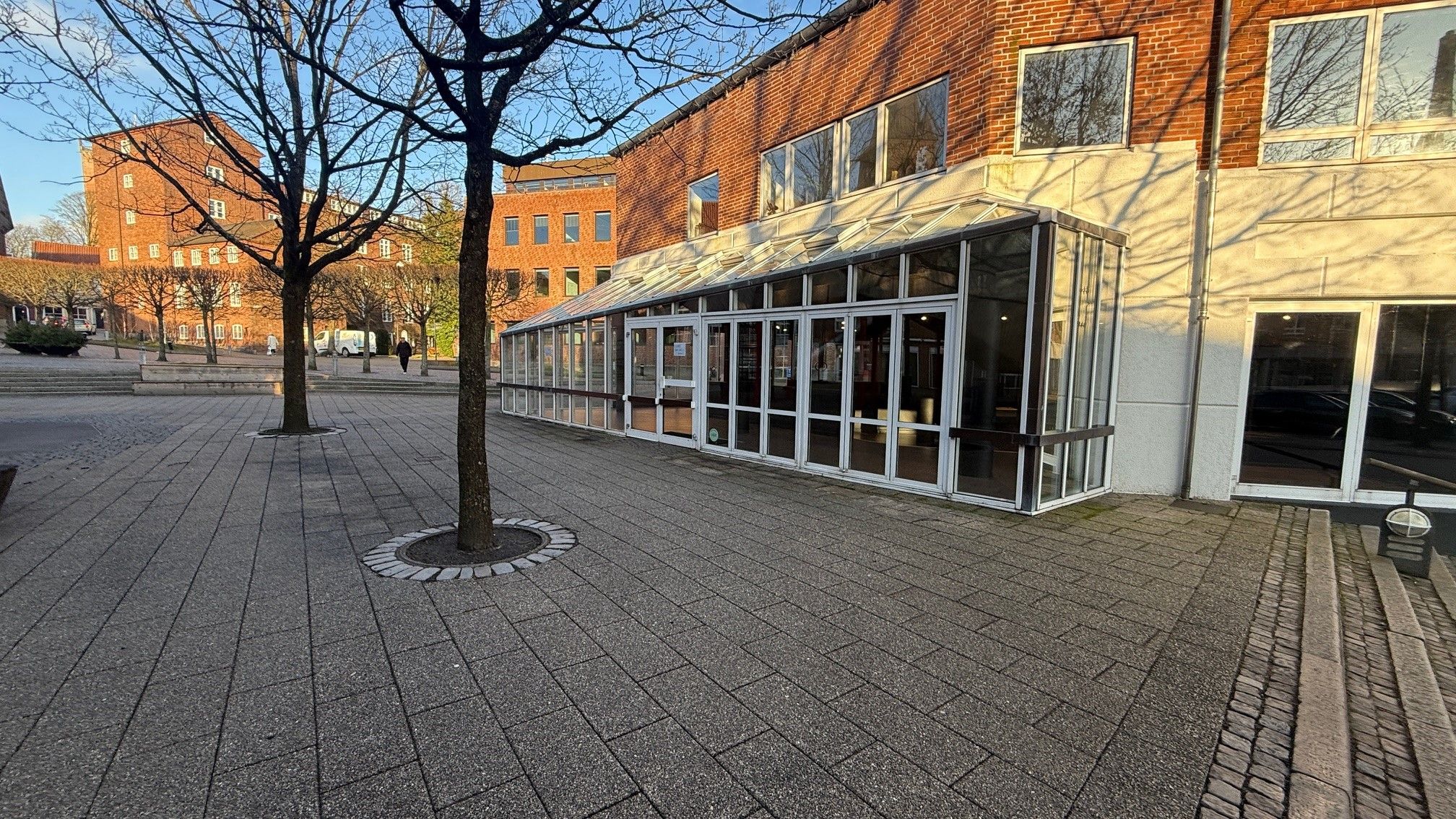 Glasveranda foran en rødmaurstensbygning med flere vinduer og træer på torvet med en person i baggrunden