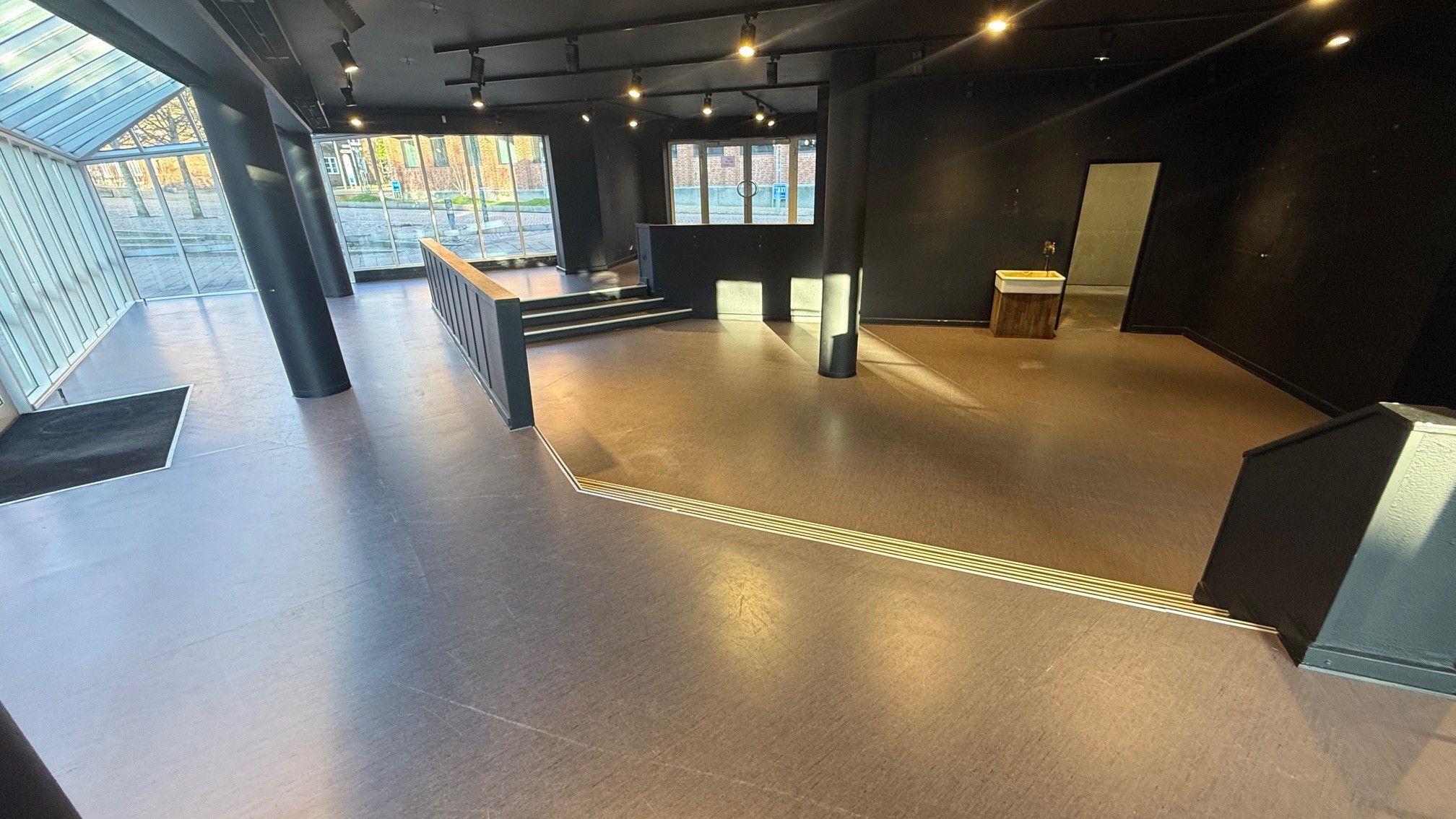 Store lokaler med lyse loftsbjælke og gul gulv. Sted med terrasse og trapper ned til en anden etage. To toiletter i højre