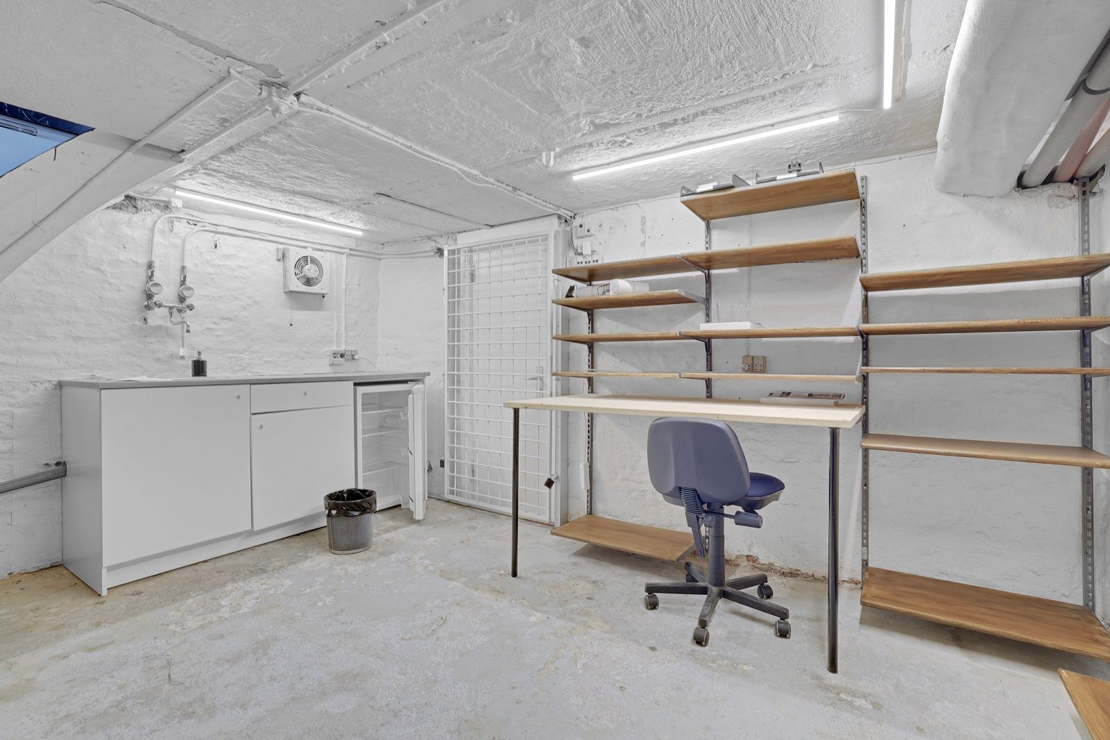 En hvid indvendig rum med loftsrum, et skrivebord, en stol og to rækker hylder. En lysekron, en ventilationsventil og en