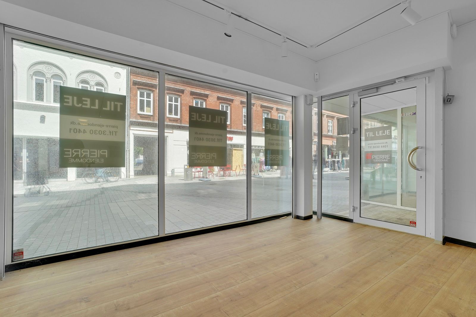 Store glasvinduer og åbne døre. Adgang til en butik med lyse, trægulve. Butikken ligger på en travl gågade med en facade mod