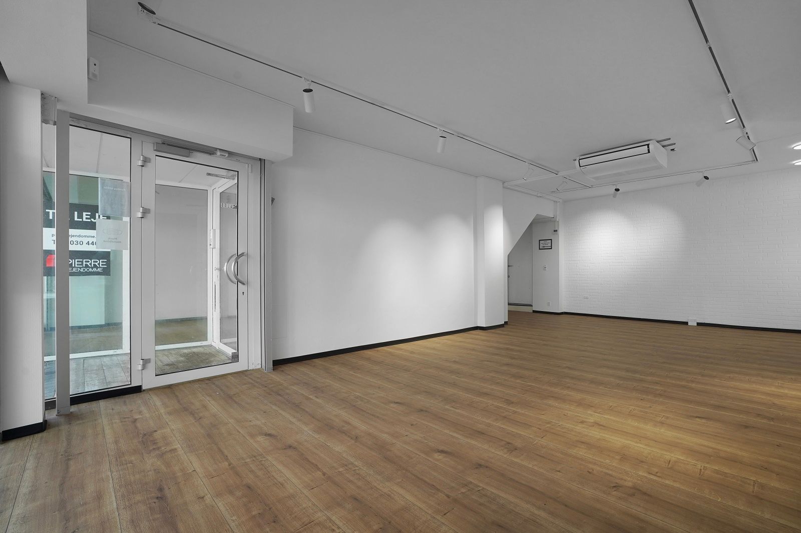 Et tomt kontorlokal med hvide vægge, trægulv og luftkanaler på loftet. Luftkanalerne og lyskarene er hvide. Der er et