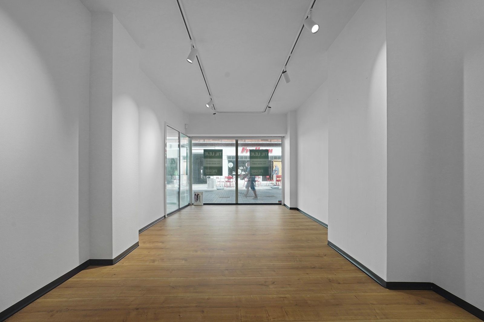 En tom butikslokal med hvide vægge og trægulv. En stor glasdør og vinduer giver lys og udsigt ud på gågaden. Loftets