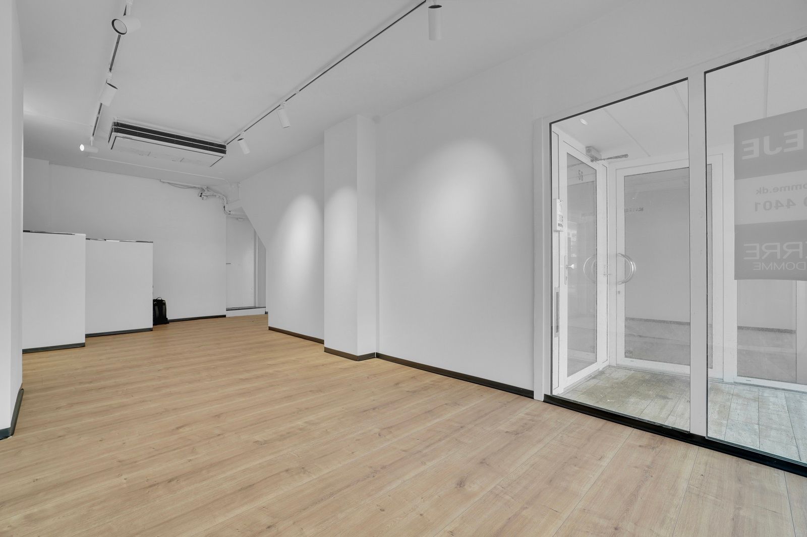 Kort række af hvide vægge og lyse trægulve i en stor forretningslokal med to store glasdøre og store vinduer. Airconditioner