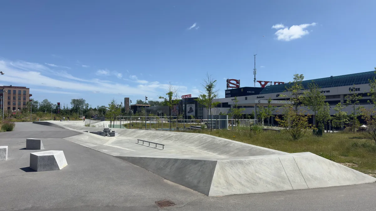 En skatepark med udfordrende ramper og grønne områder i baggrunden, hvor City og flere butikker er synlige