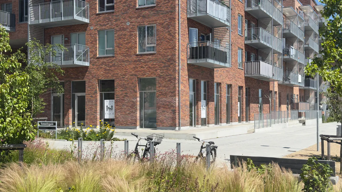 Lejlighedshus med parkerede cykler foran, grønne planter og blomster i forgrunden og et skilt med teksten Ronnebygade