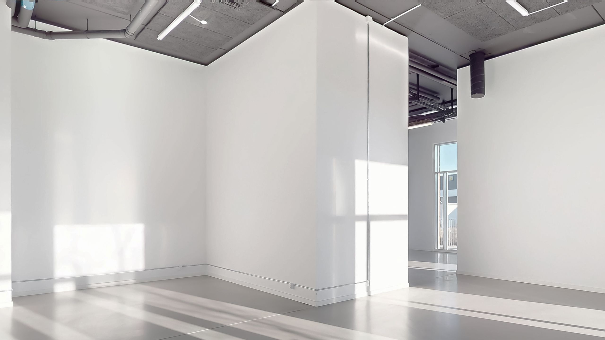 Et stort, lysafskærmet og rent værelse med hvide vægge og gulv samt en udgang til et mindre rum med glasdøre