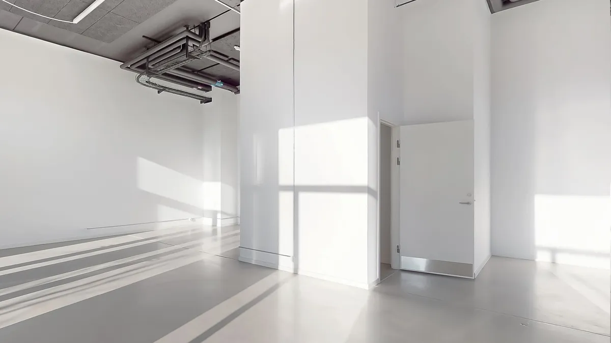 Et minimalistisk rum med hvide vægge og gulv, åbne døre og en lyse skygger på gulvet fra vinduet