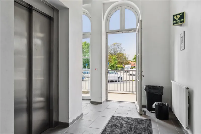 Køkken med lyseruder og trægulv samt lyse indgangsdøre og elevator samt 2 skraldespande og radiator