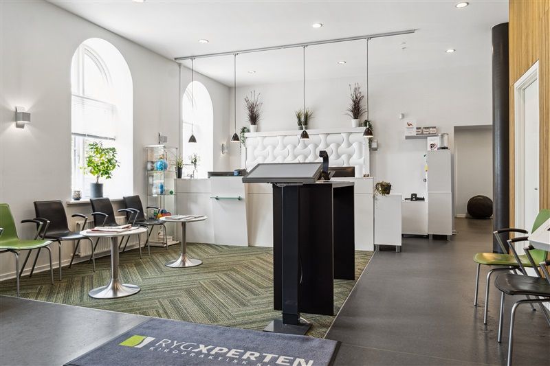 RygXperternes kontorlokaler med moderne møbler, plantehyller, stolrække og højt talerstativ foran et gulvtæppe med logo