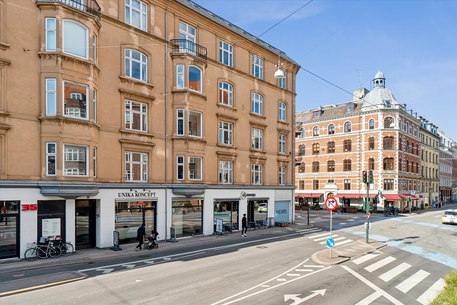 En bygning med butikker på gadeniveau, et kryds med zebraoverskridelse og et stort rødt bygning i baggrunden