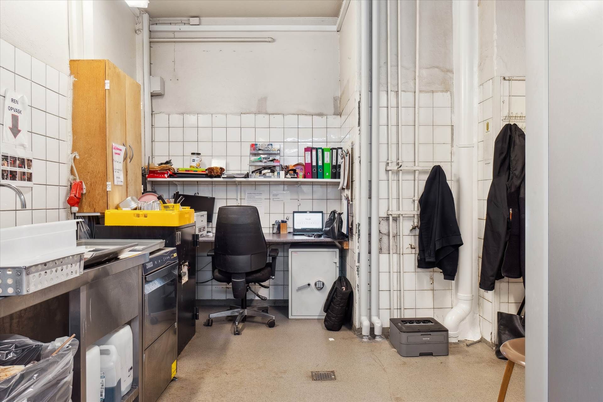 Kontor med en stor computer, skrivebord, stol, strygejern, kasseapparat, tøjstativ og flere kasseapparater i et kontorlokal.