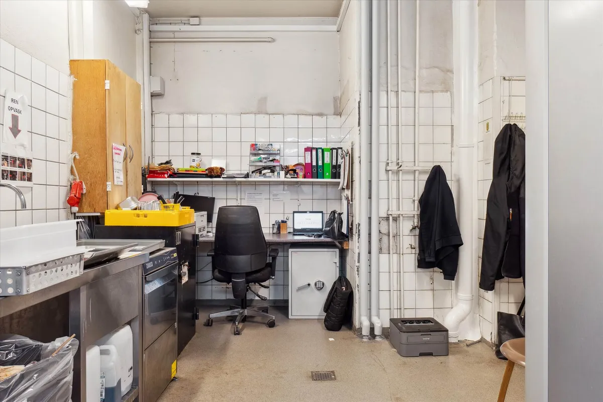 Kontor med en stor computer, skrivebord, stol, strygejern, kasseapparat, tøjstativ og flere kasseapparater i et kontorlokal.