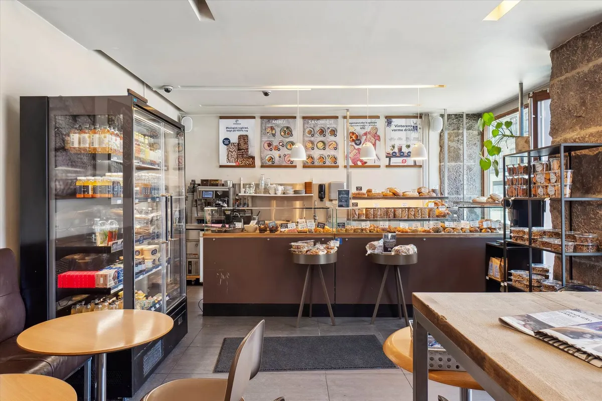 Butik med små borde, stole, køkkenudstyr, køleskabe og skuffer med slik og snacks. På bordene er der aviser og flere små