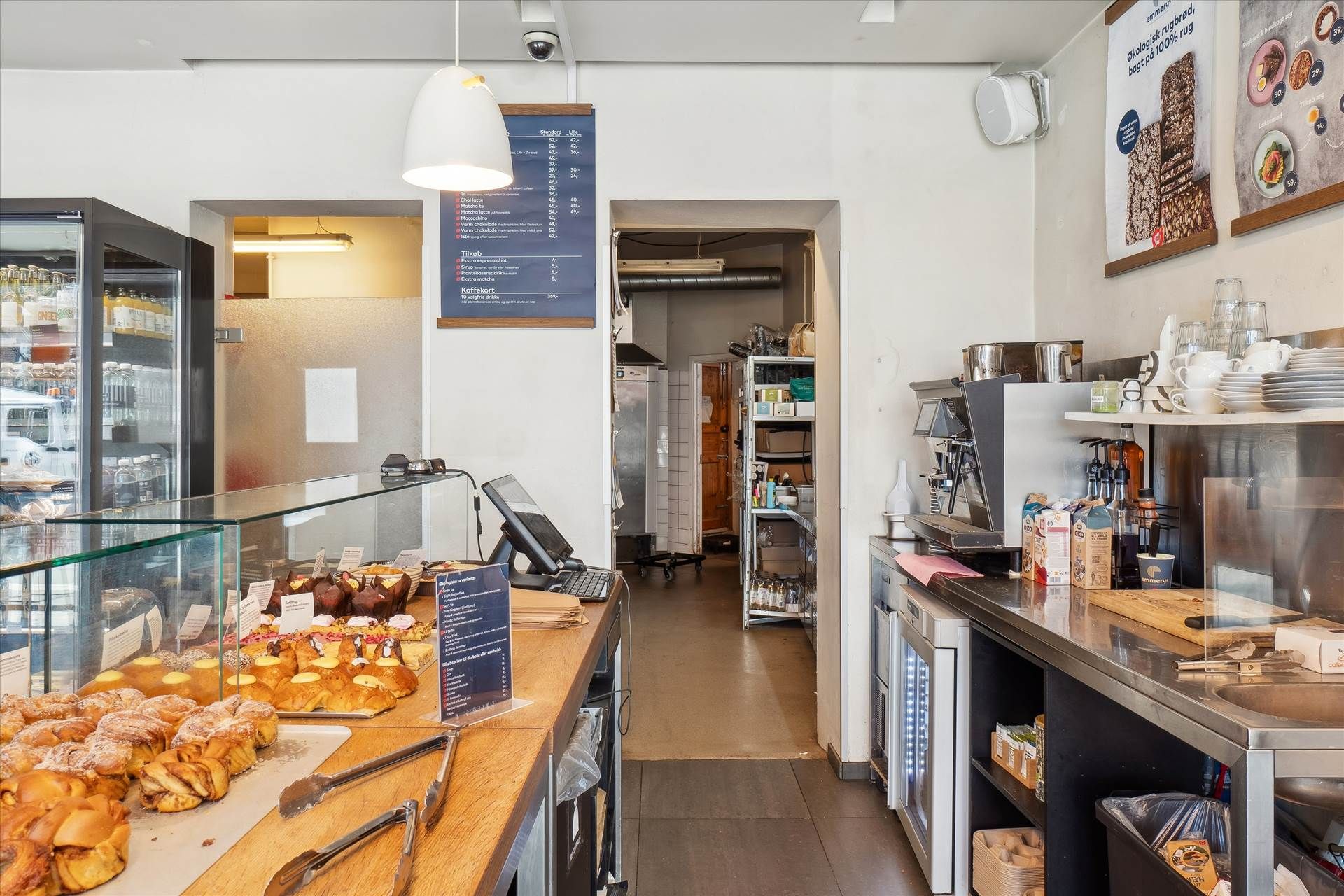 Indkøbsområde med udstillingsvindue og salgsbord, køkken med kaffemaskine og flasker, glaskøleskab med forskellige