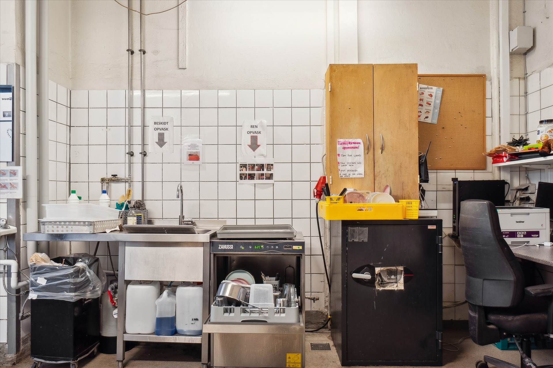 Et køkken med vask, opvaskemaskine og køleskab, der er fyldt med mad og flasker. Der er en kasse med en flaske og en knap på