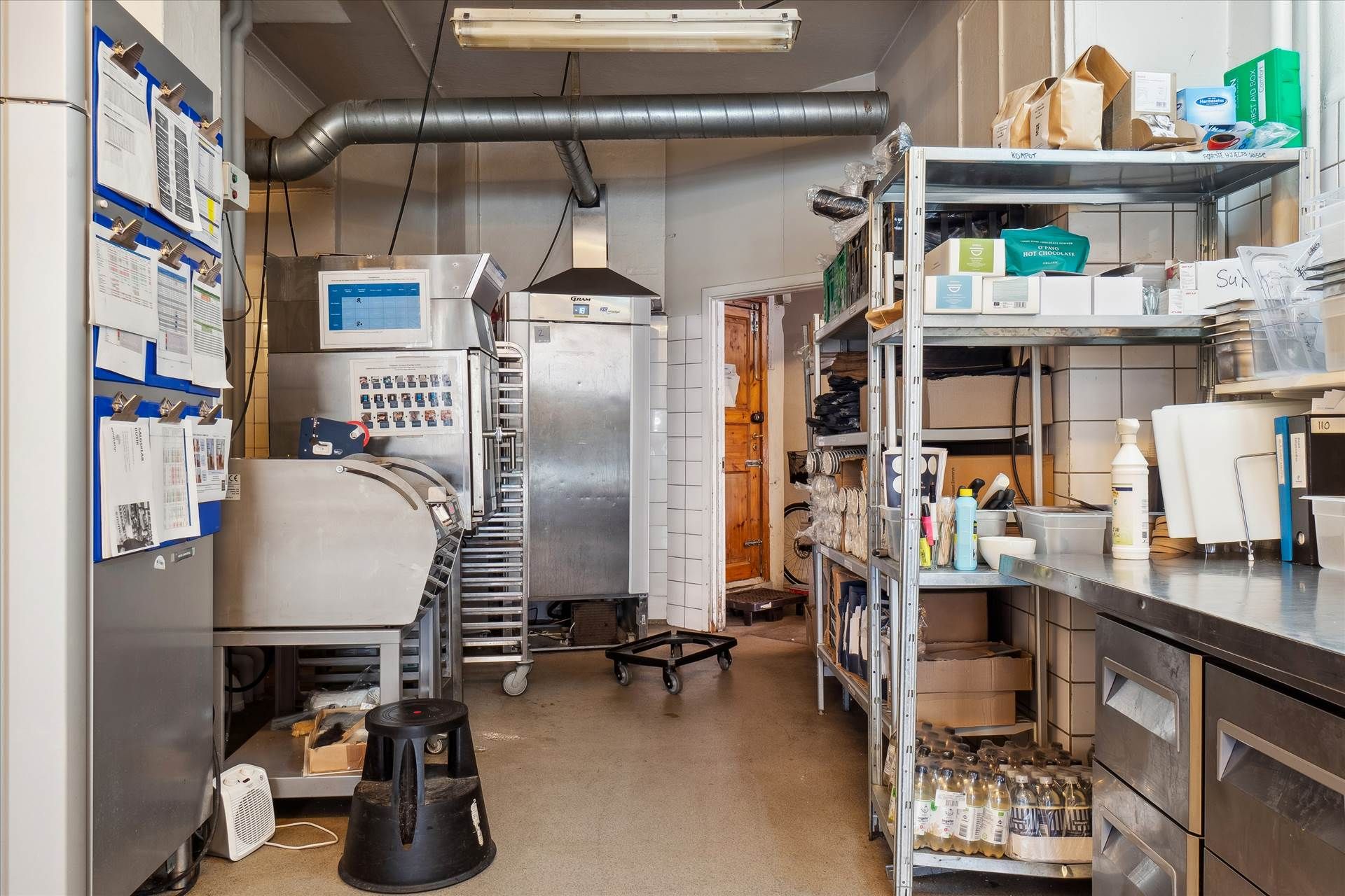 Et køkken med en stor komfur, et køleskab og flere opbevaringsskabe med papir og emballage samt en stol og et hjul under en