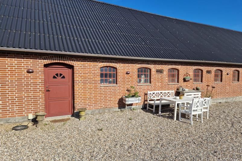 Bygning med rød dør og hvide møbler udenfor på terrassen med udsigt til sol. Planter og hængekurv på vindueskarmen.