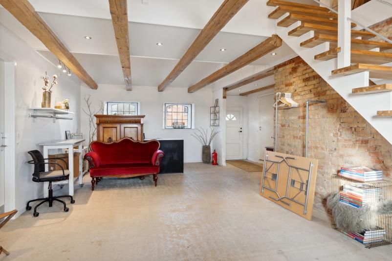 Kontorrum med lyse gulve og høje lofter. Møbler inkluderer en rødfloret sofa og en hvid bord. Stairs og møbler i loftet og