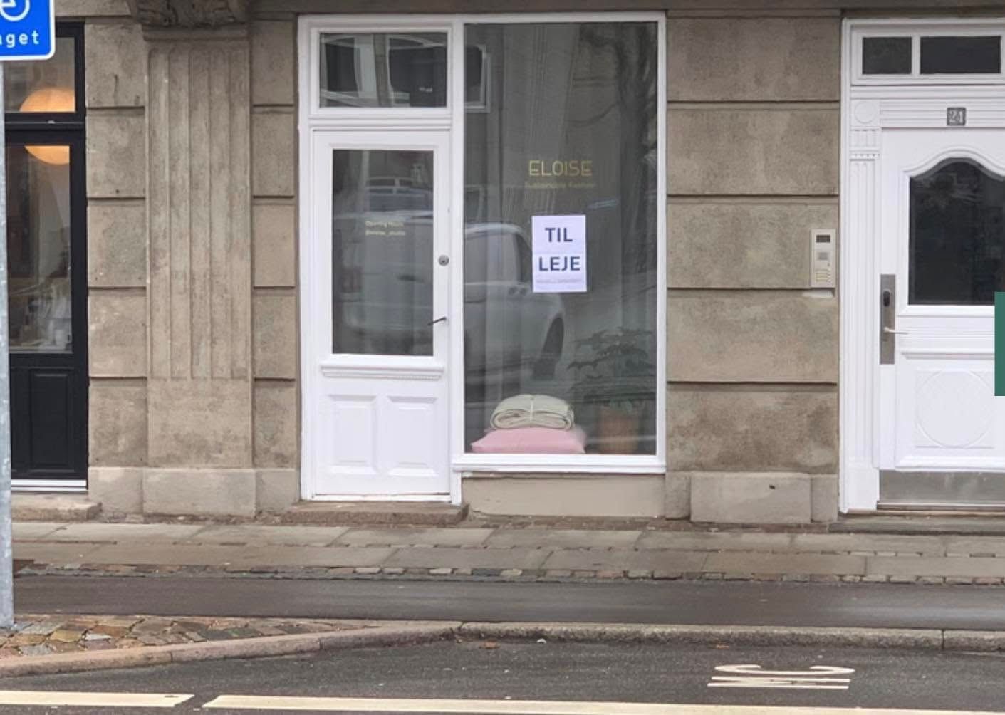 En butiksvindu med en hvid skilt med teksten Til Leje og navnet Eloise. Vinduet er lukket og reflekterer en bil og et hvidt
