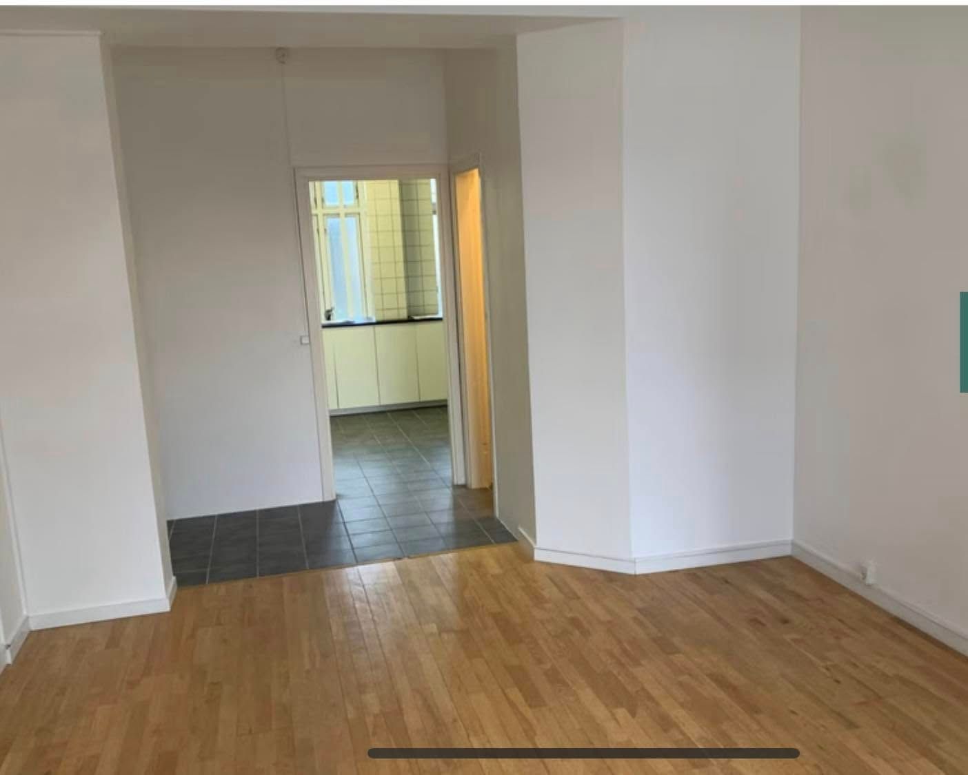 En stor og lys stue med lysegule vægge og trægulv samt et køkken og et badeværelse i baggrunden