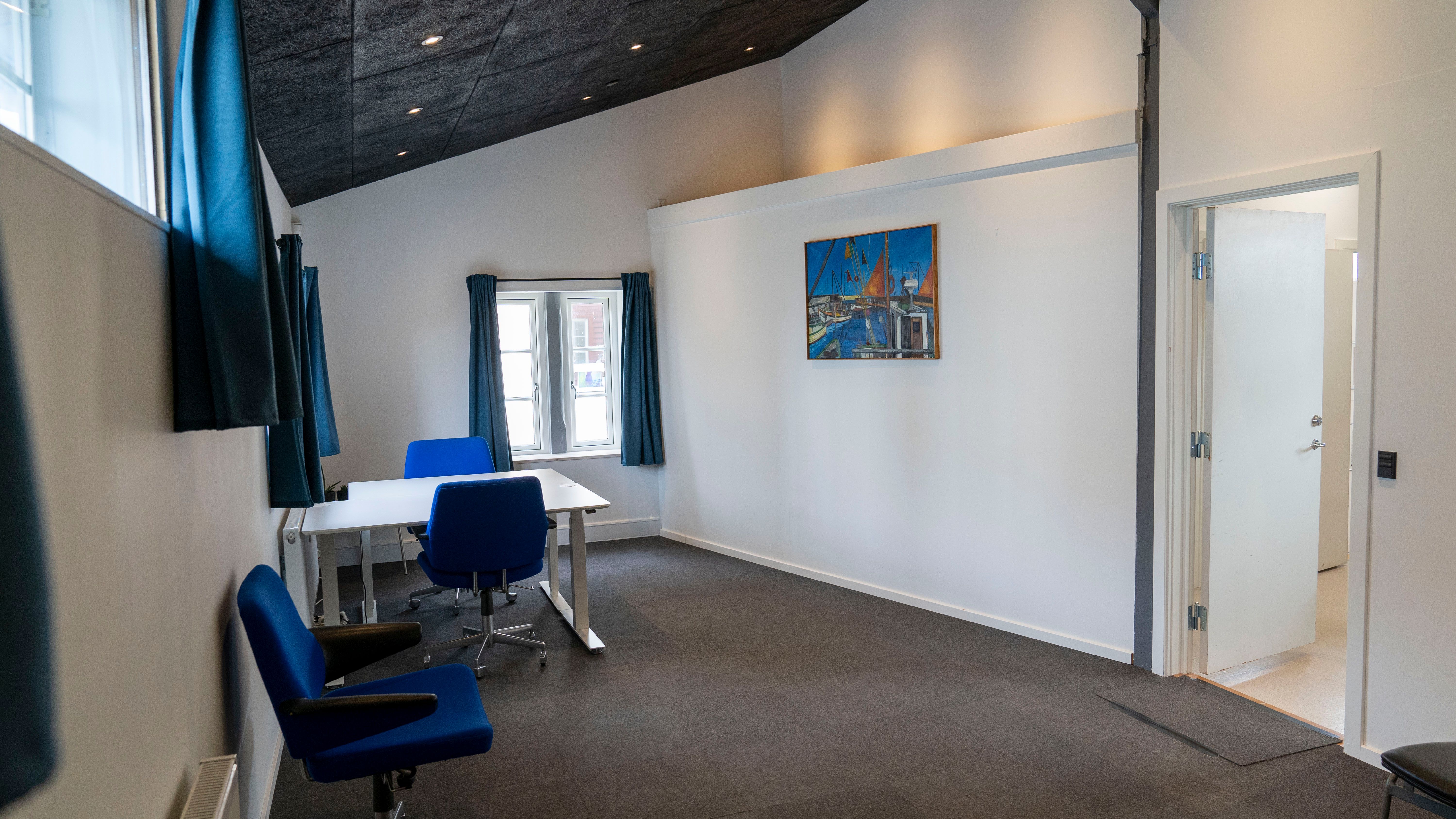 Lyst hjørnekontor med skrivebord og stole og vinduer med gardiner. Maleri på væggen og højt til loftet.