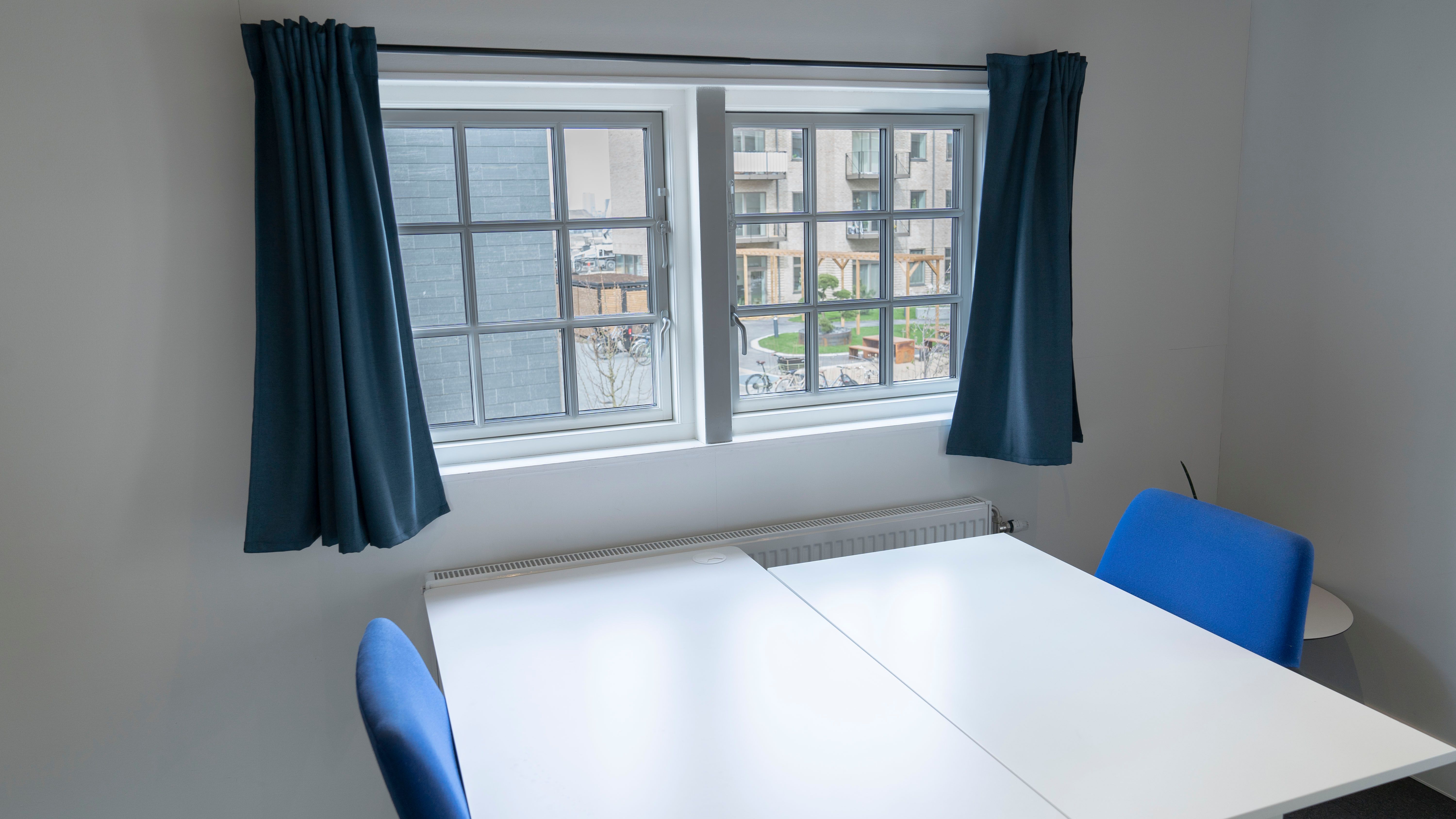 Hjørnekontor med to stole og skrivebord, vinduer med gardiner og et radiatorpanel. En gård med planter og træer er synlig