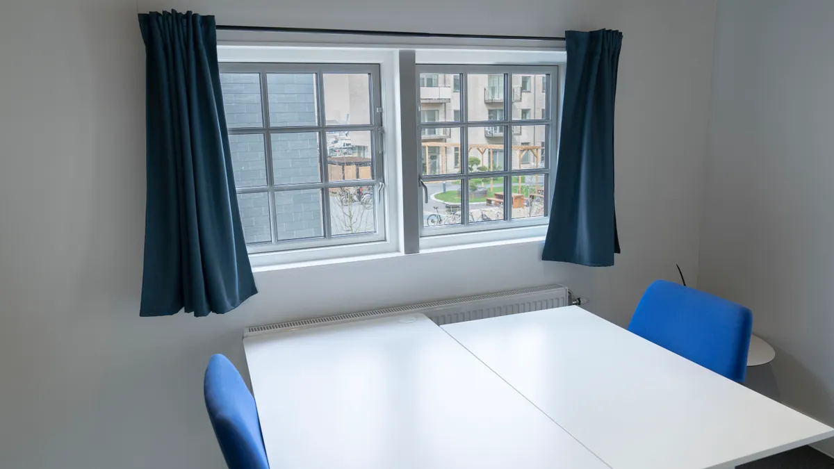 Hjørnekontor med to stole og skrivebord, vinduer med gardiner og et radiatorpanel. En gård med planter og træer er synlig